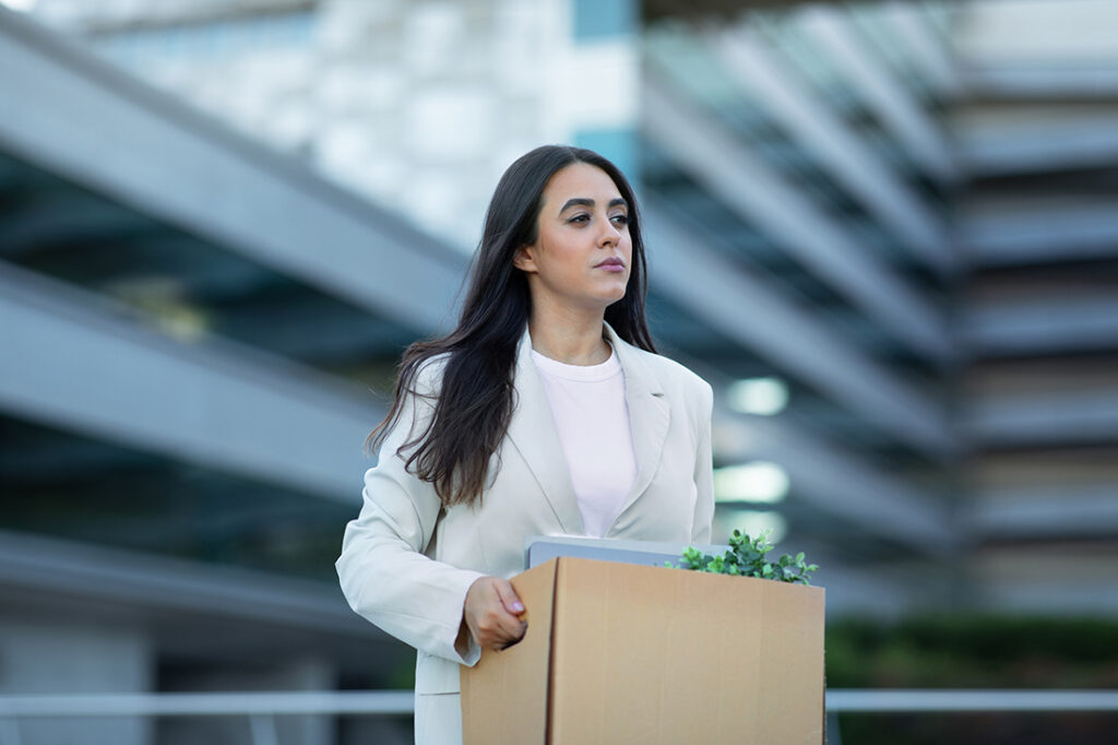 Recently laid off employee carrying cardboard box with her belongings, leaving office building after layoff, reflecting economic challenges of corporate dismissal and unemployment