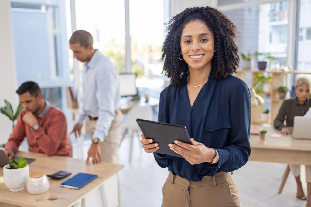 black woman holding tablet and smiling in office environment and people working in backdrop