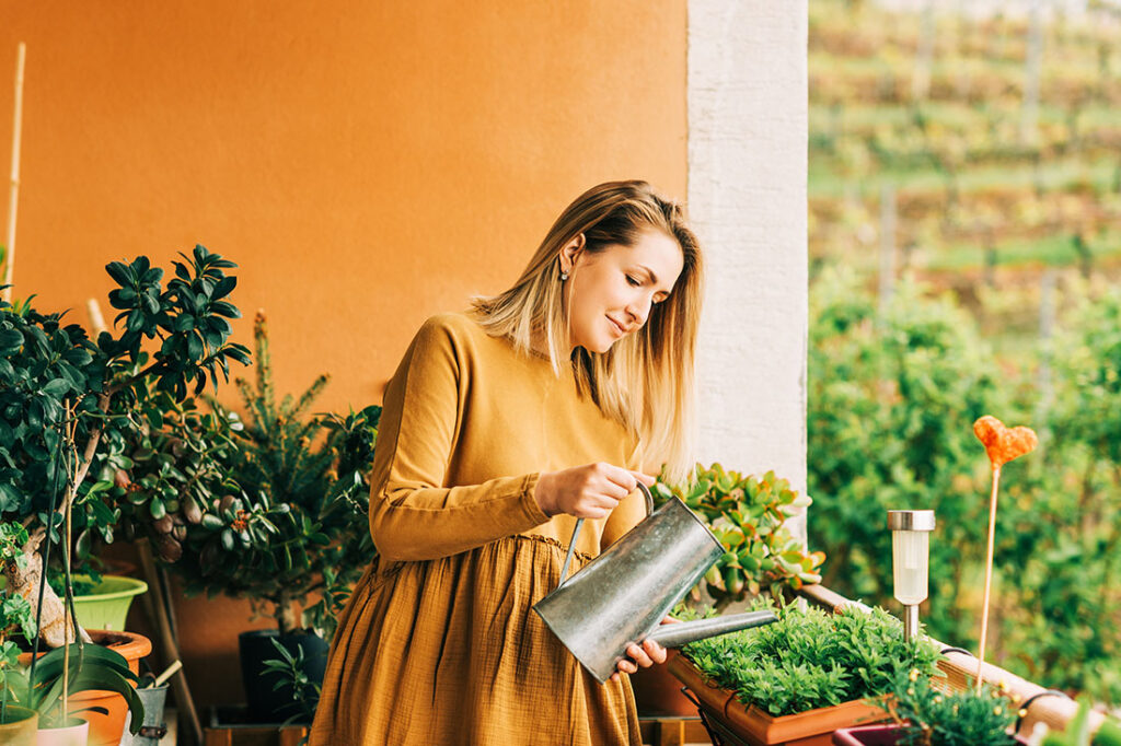 woman watering green plants on the balcony, wearing brown cotton dress
