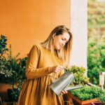 woman watering green plants on the balcony, wearing brown cotton dress
