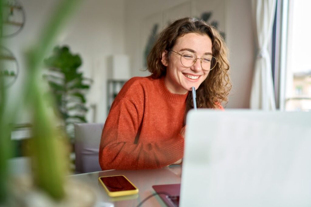 a woman sitting at her work desk while smiling at her laptop screen and holding a pen near her face