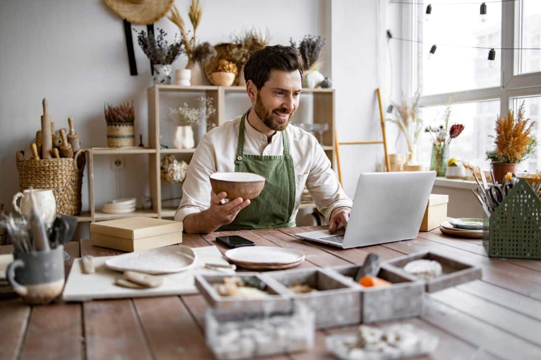 A small business owner sits at a desk in his place of business holding a bowl with one hand and using his laptop with his other hand