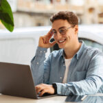 young man with curly hair and glasses smiling at his laptop in a bright room with a plant