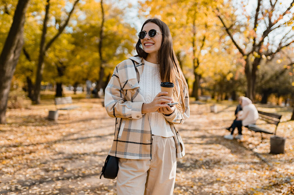 young woman walking in autumn park with coffee wearing checkered coat, sunglasses
