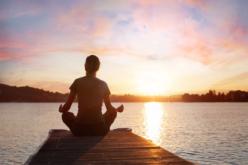 woman meditating on wooden pier near lake at sunset