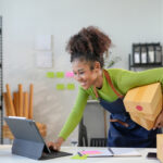 Young entrepreneur is managing her small business from her home office, smiling while carrying packages and using a digital tablet