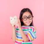 A little girl with large glasses smiles while holding up a piggy bank against a pink background.