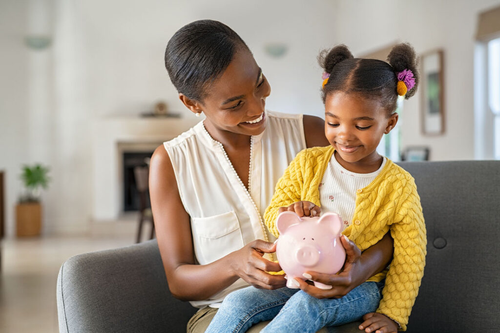 mother helping daughter sitting on lap putting money in piggy bank