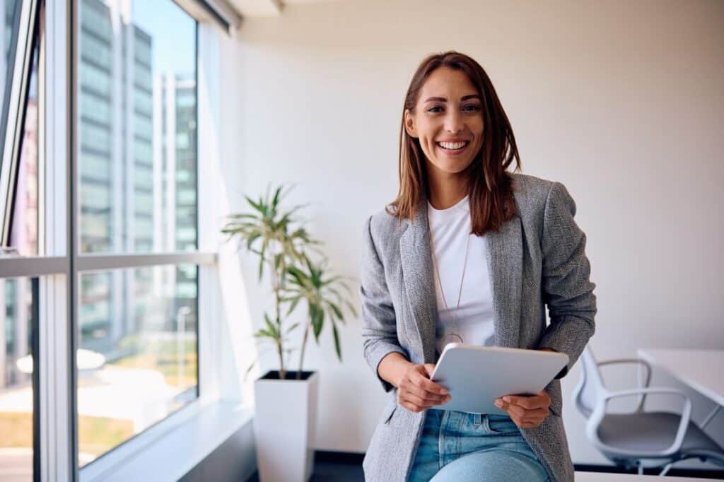 a businesswoman in an office smiling at the camera while holding a tablet device